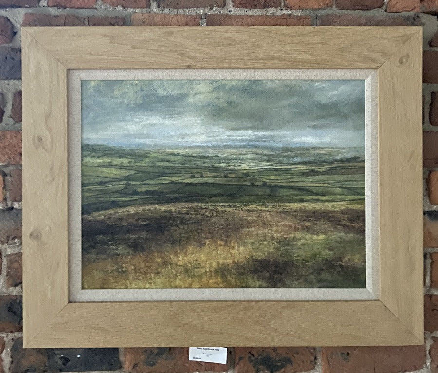 Buy 'Fields and Distant Hills', a Northern countryside landscape of patchwork fields by Sue Lawson. Image shows a painting of a patchwork field made up of greens and yellows with a field of reds and yellows in the foreground and a grey atmospheric sky above. The painting is displayed on a brick wall in an oak coloured frame with a linen mount.