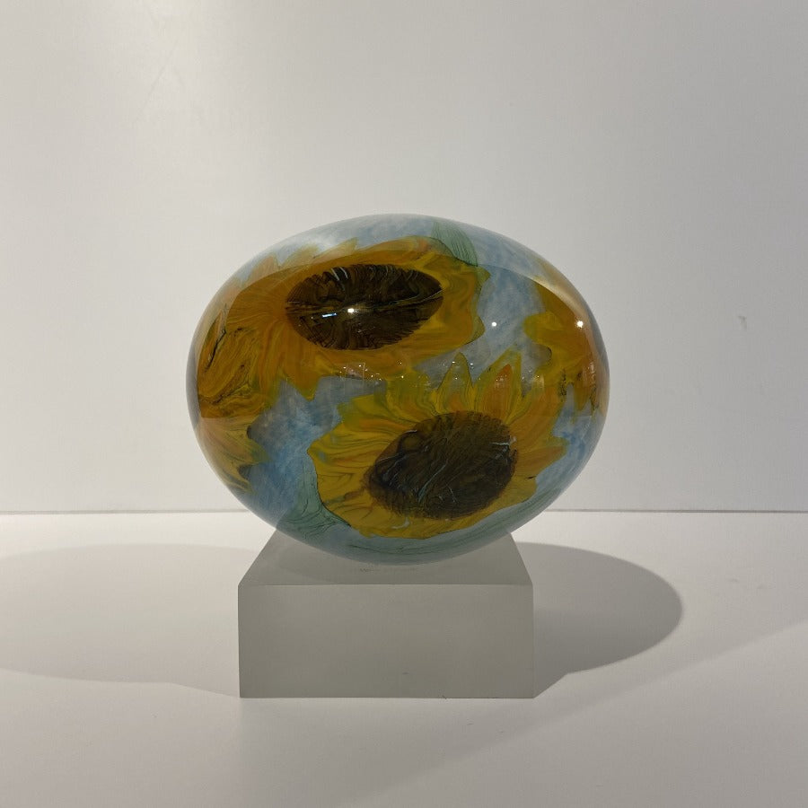Buy 'Sunflowers' blown glass by Peter Layton online at The Biscuit Factory. Image shows a round glass pot standing on a light grey block. It is light blue with several bright yellow sunflower shapes in the glass.
