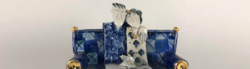 Buy handmade ceramic sculpture by Helen Martino at The Biscuit Factory. Image shows a sculpture of a man and woman sitting cuddled up on a patchwork sofa. The piece is blue and white with gold accents and the background is grey.