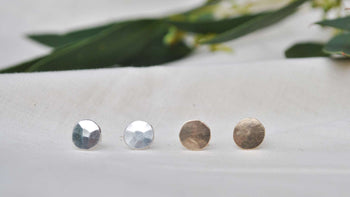 View and buy unique handmade jewellery by Sarah Ruth Stanford online at The Biscuit Factory. Image shows two pairs of stud earrings, one gold and one silver both with a bevelled surface, sat on linen with green leaves in the background.