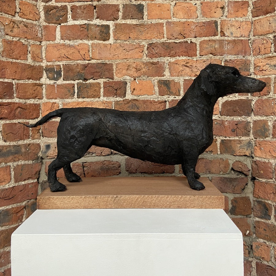 'Buy 'Sausage Dog' mixed media sculpture by Zoe Robinson. Image shows a mixed media sculpture of a sausage dog. The dog is black and is displayed on a plinth against a brick wall.'