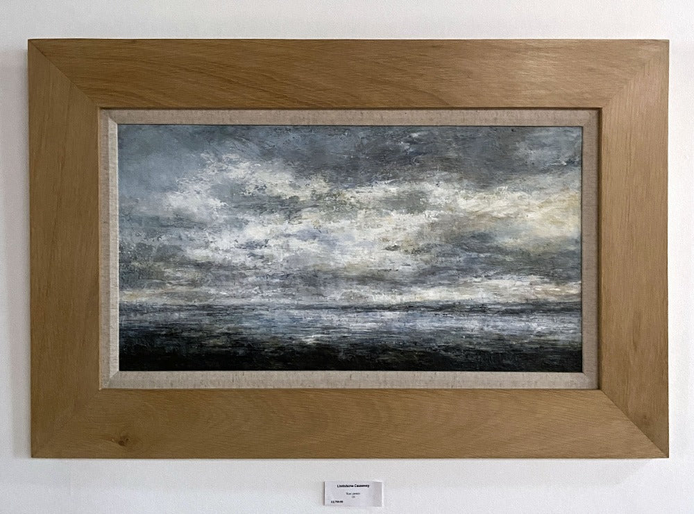 Buy Buy 'Lindisfarne Causeway at low tide Northumberland', an atmospheric landscape by Sue Lawson. Image shows a painting of a coastal scene in stormy blues and greys with pale yellow highlights. The painting is displayed on a white wall in an oak coloured frame and with a linen mount.