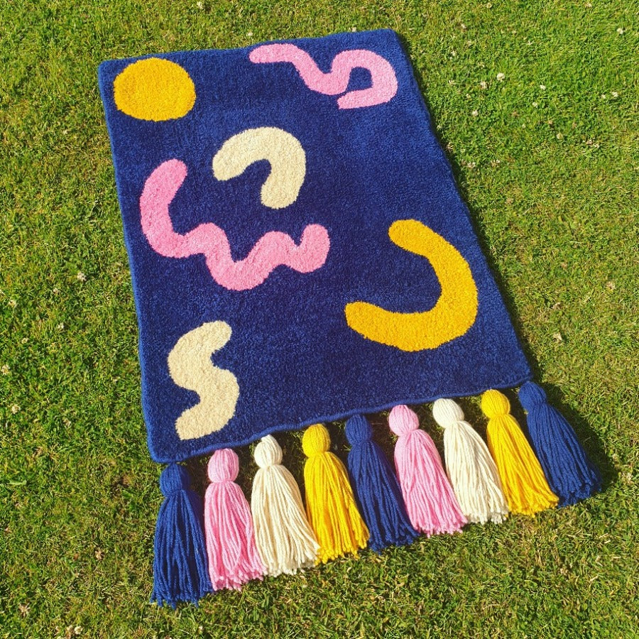 'Joy' by textile designer Loop and Yarn. Image shows  a dark blue rug with cream, yellow and pink shapes. and tassels on one edge.