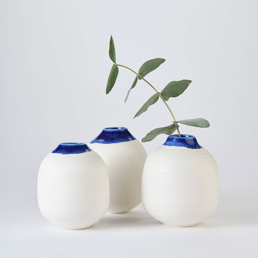 Buy 'Small White and Cobalt Moon Jar', a rounded ceramic vessel by Kirsty Adams. Image shows three small rounded white pots with a short, slightly fluted neck coloured in a vivid blue glaze. The pots form a triangle and are shot front on. In the leftmost pot a stem of green leaves sprouts from inside. The vases sit on a pale grey background.