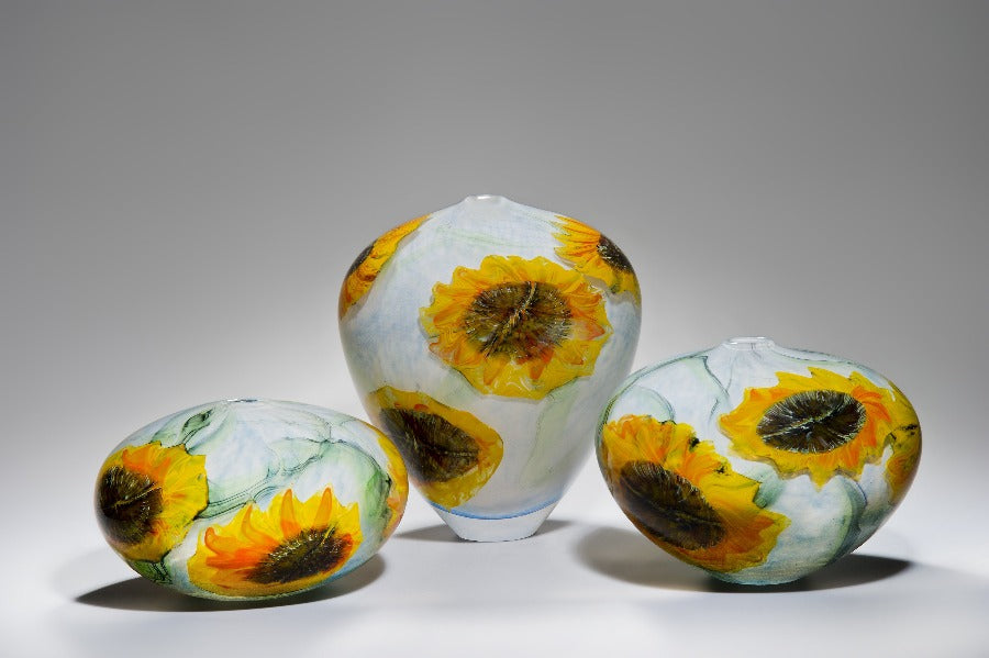 Buy 'Sunflowers' blown glass by Peter Layton online at The Biscuit Factory. Image shows a collection of three glass pots in different shapes and sizes. They are light blue with bright yellow sunflower shapes in the glass.