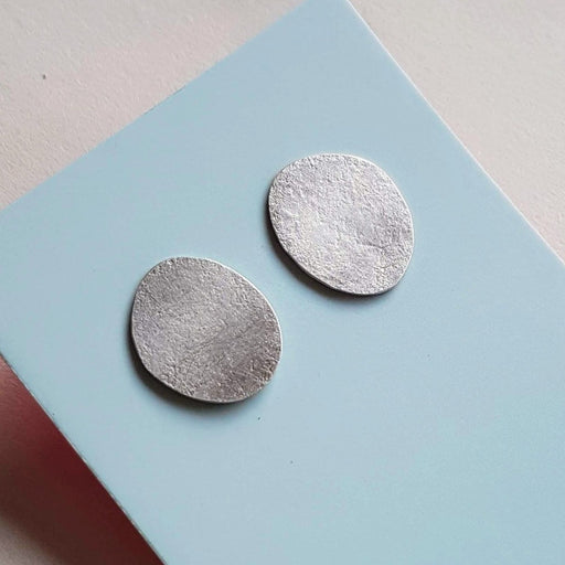 Image shows 'Lichen Earrings', handmade jewellery by Tina MacLeod for sale at The Biscuit Factory. Two irregular oval silver earrings against blue card.