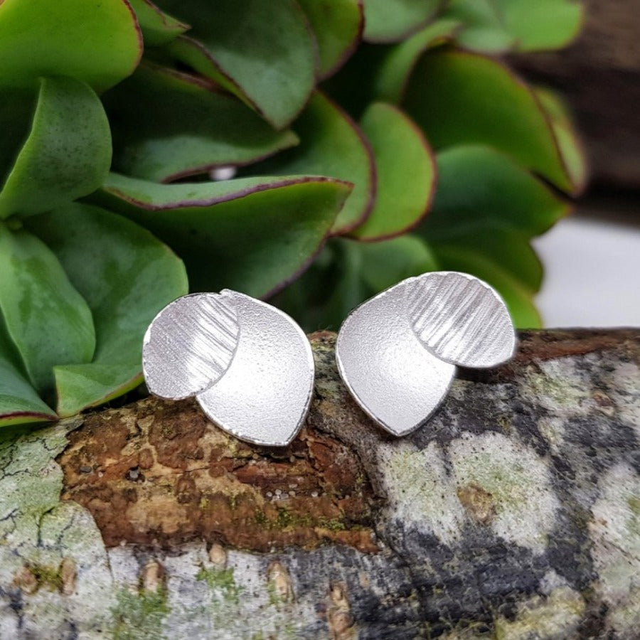 View and buy ethical handmade jewellery online at The Biscuit Factory. 'Orchid Leaf Earrings' silver floral studs by Donna Barry. Image shows a pair of earrings made up of a pointed oval overlapped by a small textured pointed oval. The earrings sit on a natural wooden plinth with green foliage in the background.