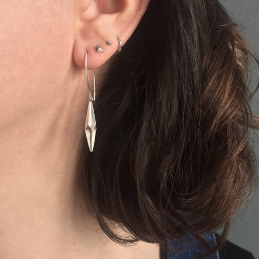 Shard Drop Earrings by Alice Barnes contemporary jewellery for sale at The Biscuit Factory art gallery. Image shows a person with brown hair wearing a silver earring in an elongated diamond shape on a silver hoop.