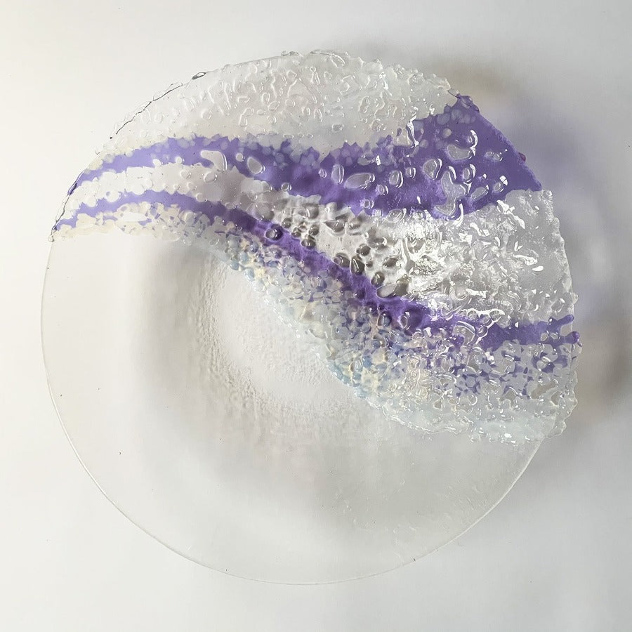 Buy 'Authenticity' a handmade decorative glass dish by Stevie Davies. Image shows a transparent glass dish decorated with fused glass details both transparent and purple in wavy lines across the surface of one half. The dish sits on a white background.