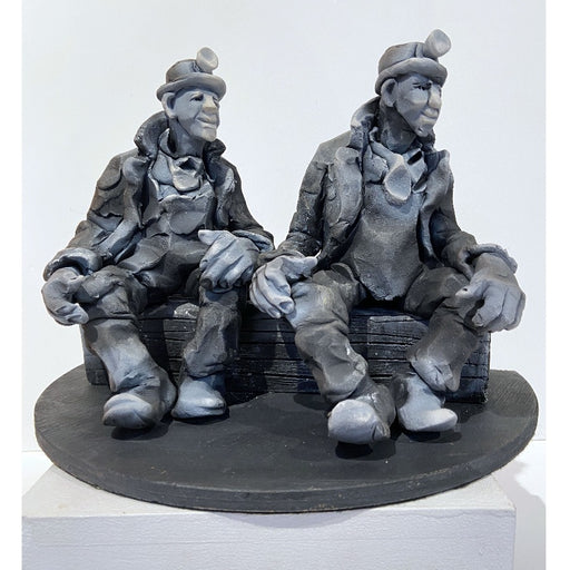 Buy 'Summints Up', a ceramic sculpture of miners by Yorkshire artist Alistair Brookes. Image shows a stylised grey sculpture of two miners wearing headlamps, large coast and boots sat on a long log both looking to the right. The sculpture is sat on a white plinth against a white wall.