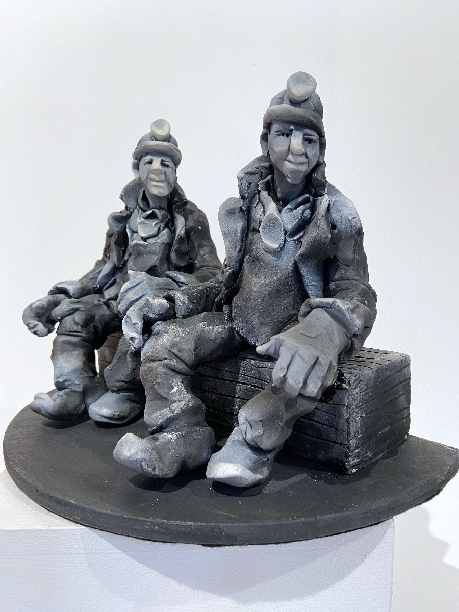 Buy 'Summints Up', a ceramic sculpture of miners by Yorkshire artist Alistair Brookes. Image shows a stylised grey sculpture of two miners wearing headlamps, large coast and boots sat on a long log both looking to the right. The sculpture is sat on a white plinth against a white wall.