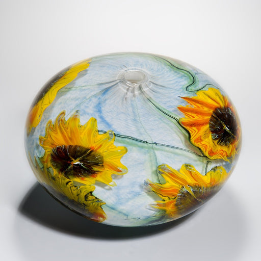 Buy 'Sunflowers' blown glass by Peter Layton online at The Biscuit Factory. Image shows a round glass pot with a small round opening in the centre of the top. It is light blue with two bright yellow sunflower shapes in the glass.