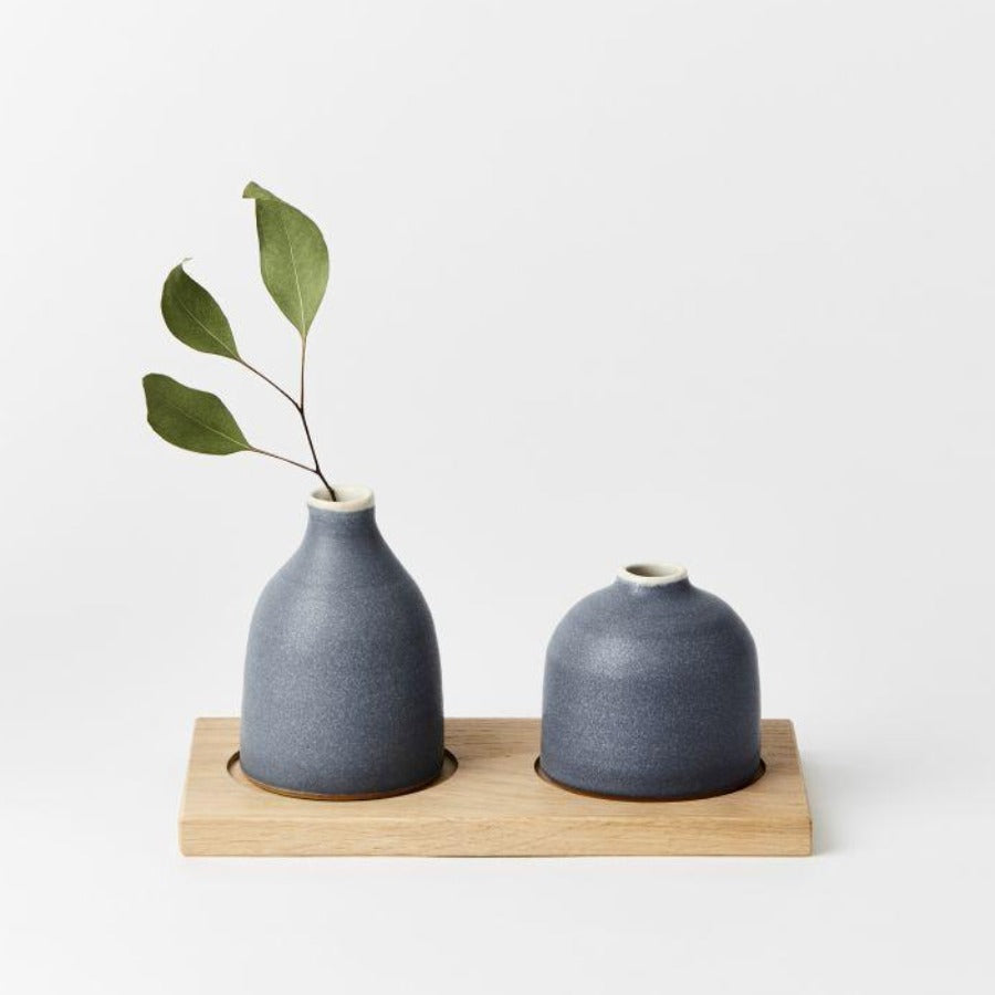 Buy Duo of Bud Vases on Oak Board, original handmade ceramics by Tone von Krogh online at The Biscuit Factory. Image shows a pair of grey/blue ceramic bud vases on an oak board. One is slightly taller than the other and has a sprig of foliage protruding.