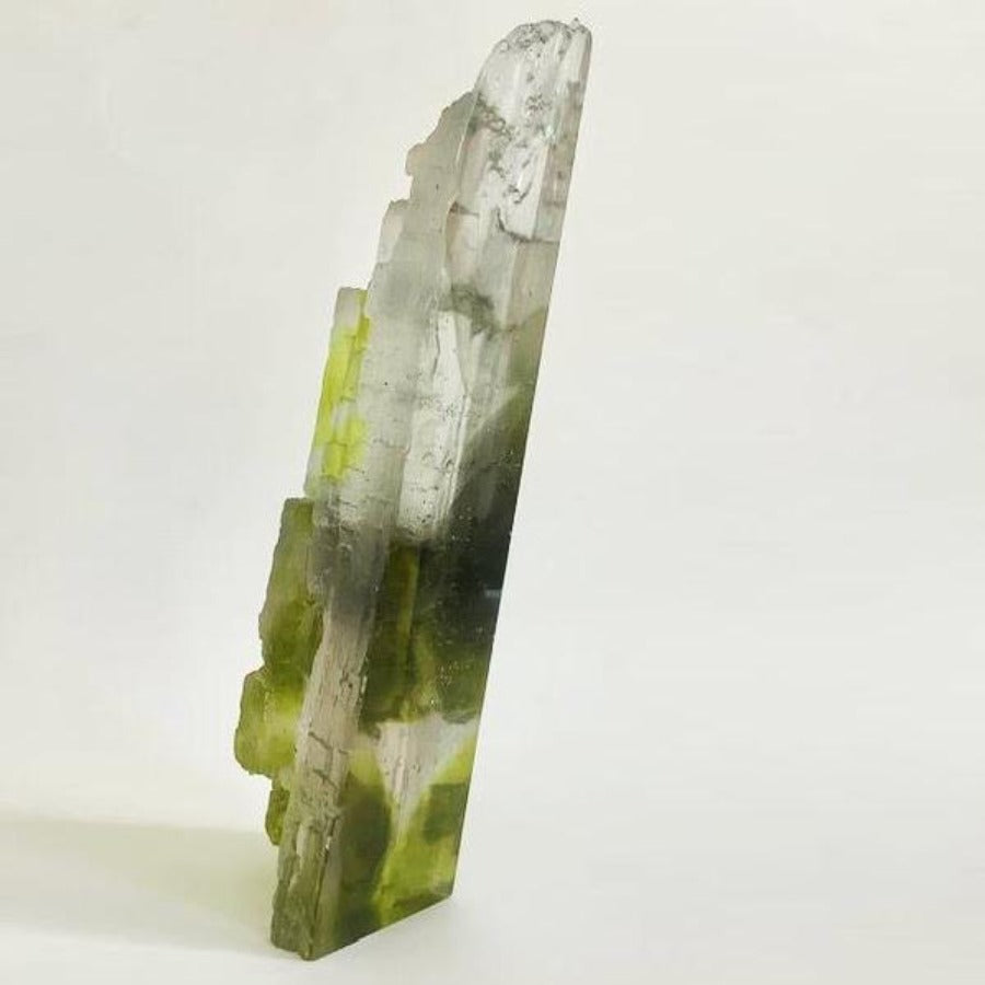Buy 'Cliff Edge' a handmade glass sculpture by Pat Marvell. Image shows a tall cast clear glass sculpture with light and dark green colouring rising up from the base. The sculpture leans slightly to the right and is sat on a beige background.