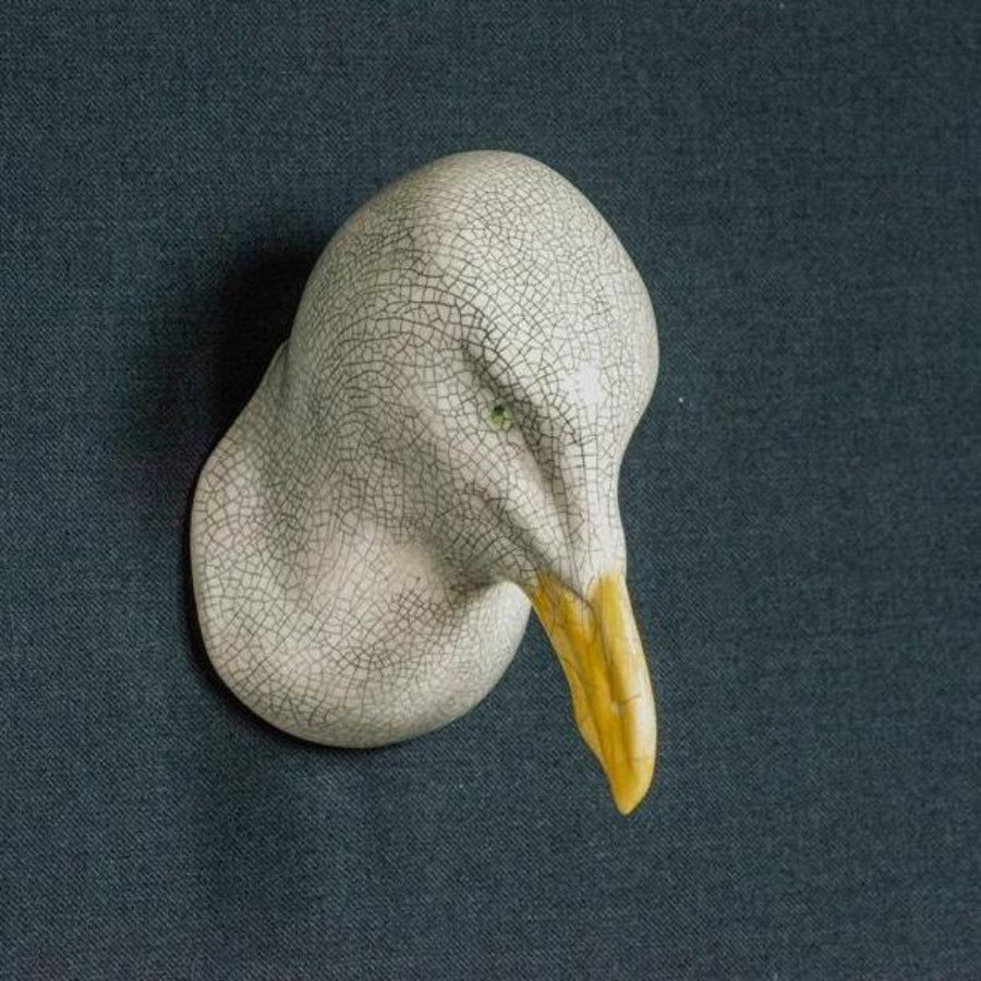 Buy handmade ceramic sculpture by Jackie Summerfield online at the Biscuit Factory. Image shows a ceramic sculpture of a seagulls head against a dark blue background. The sculpture is white with a crackled glaze. The seagull has green eyes and a yellow beak.