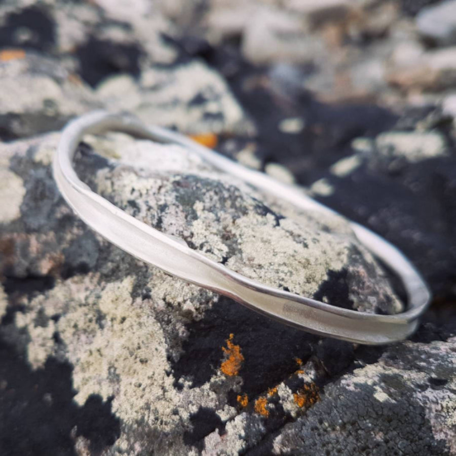 Buy 'Mara Bangle', handmade jewellery by Tina MacLeod at The Biscuit Factory, Newcastle upon Tyne. Image shows an irregular shaped silver bangle on a black rock with white and yellow lichen