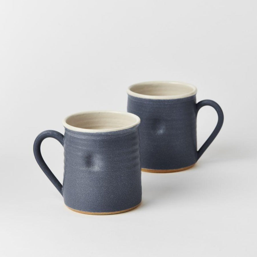 Buy ceramic mug, original handmade craft by Tone von Krogh online at The Biscuit Factory. Image shows a pair of blue/grey mugs with cream rims and interiors, standing one behind the other. They each have a dimple in the front.