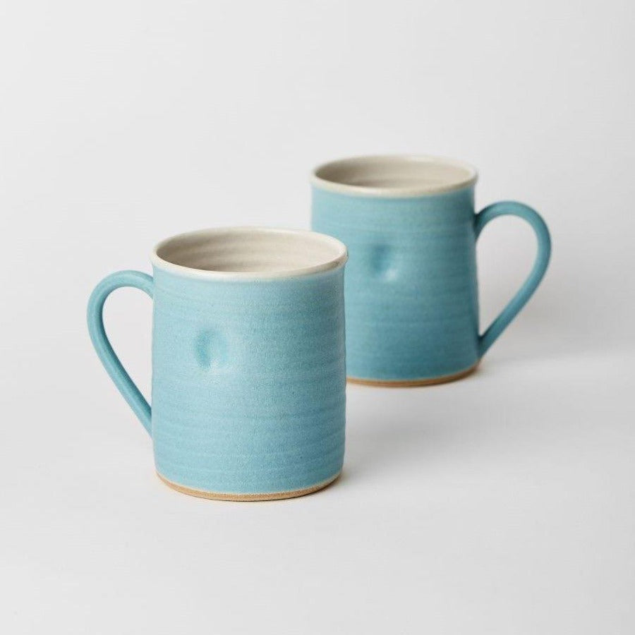 Buy ceramic mug, original handmade craft by Tone von Krogh online at The Biscuit Factory. Image shows a pair of turquise mugs with cream rims and interiors, standing one behind the other. They each have a dimple in the front.