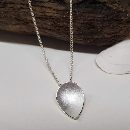 View and buy ethical handmade jewellery online at The Biscuit Factory. 'Plain Pyrus Pendant' silver floral necklace by Donna Barry. Image shows a silver pointed oval pendant on a white background attached to a chain which lays over a piece of wood at the top section of the frame.